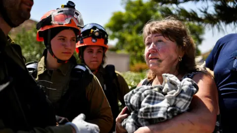 Reuters Een vrouw praat met Israëlische eerstehulpverleners nadat haar huis werd beschadigd door raketvuur van Hezbollah in Katzrin, op de door Israël bezette Golanhoogten (21 augustus 2024).