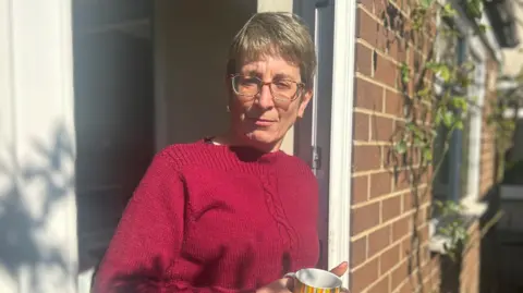 Christine Bell in magenta knitwear standing at a door holding a striped mug. Christine is in her 50s with cropped dark grey hair and wearing glasses.
