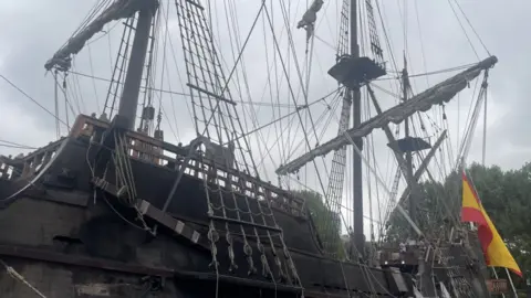 A close-up shot of a full-scale 17th Century Spanish tall ship, made of wood with riggings and curled up sails. There is a yellow and red flag hanging from one of the ropes.