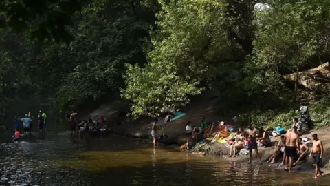 Getty Images A group of people are gathered by a riverbank surrounded by lush green trees. Some individuals are wading in the water, while others are sitting or lying on towels on the sloping bank, relaxing and socializing. The scene is shaded by overhanging branches.