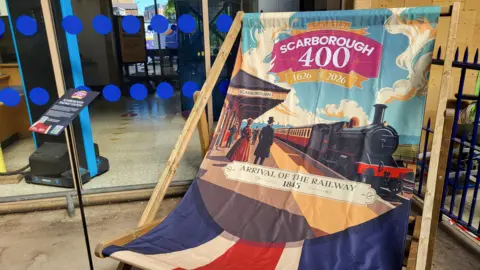 A giant decorative deck chair stands outside a building with glass doors, its fabric printed with colourful artwork celebrating Scarborough’s 400th anniversary. The design shows a steam locomotive arriving at a station with people in period clothing, alongside text marking historical dates.