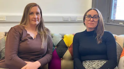 Two women sitting on a sofa and looking towards the camera. Donna, on the left, wears a brown v-neck top with a scalloped edge. Rachael, on the right, wears black glasses, and a black high-neck top and a black and white patterned skirt. The sofa has a colourful diamond pattern.