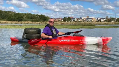 Bruce Langley in a red kayak. He is wearing a purple T-shirt and holding the paddle. He has sunglasses on and has two tyres on the back of the canoe.