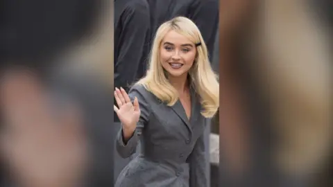 Sabrina Carpenter has long dyed blonde hair. She is wearing a tailored grey suit jacket and waving with her right hand.