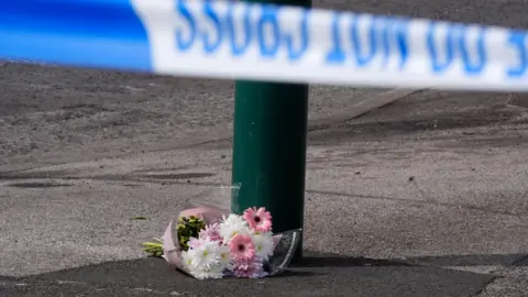 PA Media A floral tribute left near the scene, where a murder investigation has been launched after two people died in a house fire at a property in Grangetown, near Middlesbrough. A bouquet of pink and white flowers lies next to a lamppost, with a police tape in front of it