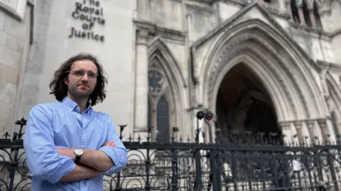 BBC/ Hazel Shearing David Hamon has shoulder-length brown hair, is wearing glasses and a light blue shirt with the collar unbuttoned and has his arms crossed as he stands in front of the Royal Courts of Justice in London