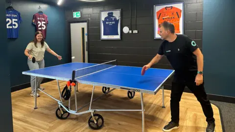 Alice Cunningham/BBC Mr Hamilton plays table tennis with one of his staff members in the Halo officers. Framed Ipswich Town shirts have been hung on the wall behind the ping pong table.