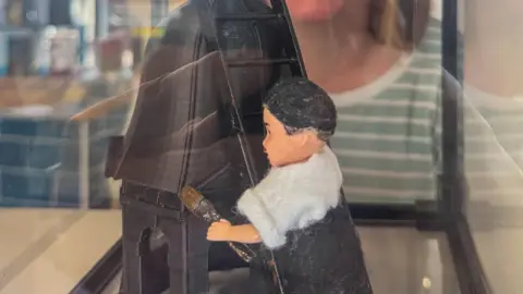 A small doll on a ladder holding a paintbrush leaning against the representation of a tower roof.