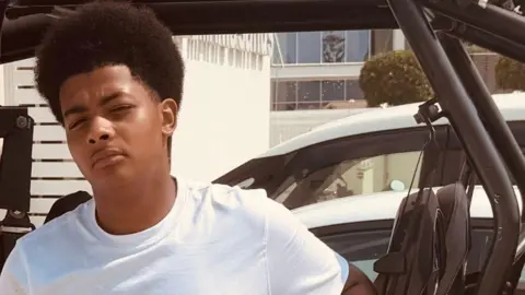 A teenage boy leaning against a car, wearing a white top