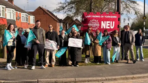 A group of people standing on a path holding up colourful signs and banners. A large red banner is behind them. It reads "East Riding of Yorkshire NEU".