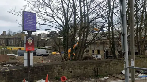 Google A tall blue signpost bearing the name Crossflatts Cricket Club in front of a building with scaffolding around it