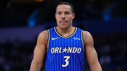 Desmond Bane seen from waist up in the centre of the image looks on in a blue Orlando Magic vest with white vertical pinstripes. A white number three is in the centre of his chest and above it the word 'Orlando' in white, with the letter A replaced by a grey five-pointed star