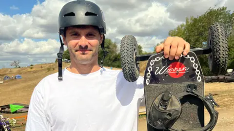 Amon holds up his board and smiles for the camera. He wears a white t-shirt and a black helmet, and his board is black with large rugged wheels and foot straps. 