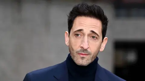 A headshot portrait of Adrien Brody taken at a photocall for The Brutalist. He is wearing a dark turtleneck jumper and navy blue jacket.