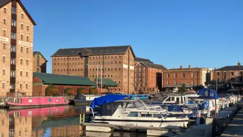 A photo of Gloucester Docks. There are a number of large orange brick warehouse buildings. In the docks there are a number of small blue and white speedboats moored up, and several canal boats behind them.