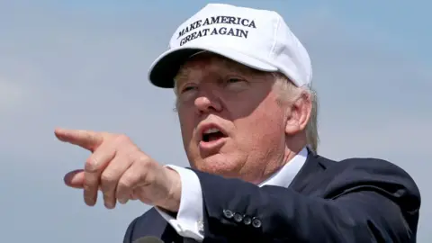 PA Media A man with a white cap points with his left hand. He is outside, with a blue sky in the background, and is wearing a navy suit and white shirt. It says "make America great again" in capital letters on his hat.