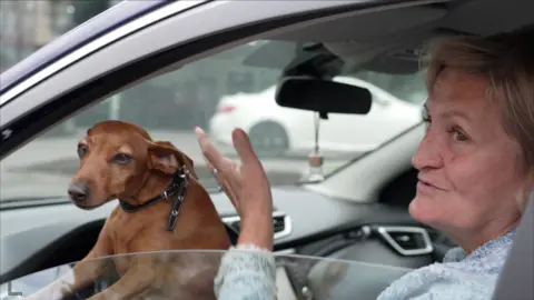 A small brown dog with a black collar peers out of a car window. It's sat on her owner's lap. Lyubov, the driver and dog owner, is wearing a blue jumper and gesturing out of the open window to the camera.