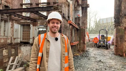 A man in a beige jacket, along with a orange hi-vis jacket and a white hard hat. He's stood in front of the remains of a building, with a dumper truck in the background.