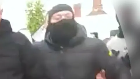 Essex Police Luke Fleming wearing a black face covering and hooded jacket. He is among a crowd of people by a police cordon.