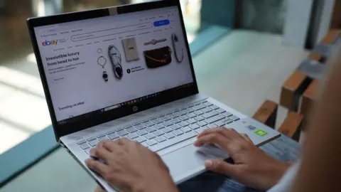 A woman browses Ebay on a laptop. Only her hands and the front page of the website can be seen. 