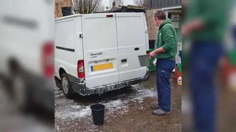 A man in a green hoodie and blue tracksuit bottoms cleaning his white van 