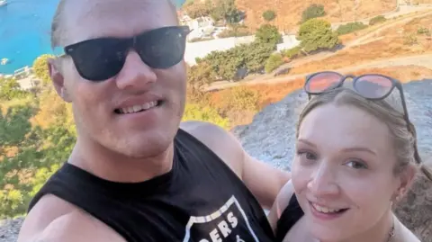 Jess - a short blonde woman in her 20s - stands to the right of her partner Jake who is wearing a black tank top and has his arm around Jess. They both are looking upwards to the camera and smiling. They're standing on a hill with a Mediterranean-looking coastal background behind them in this holiday snap. 