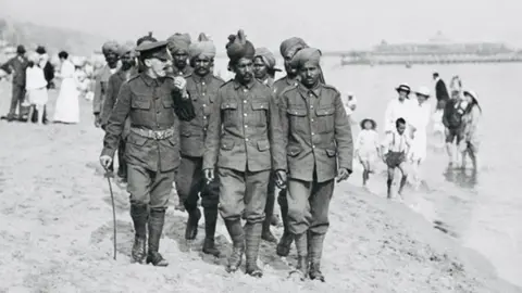 A group of Indian soldiers in uniform walking across beach with pier in background and people padding in the sea