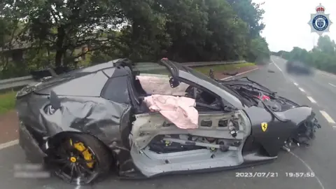 A catastrophically damaged gun-metal grey Ferrari sitting on a slip road next to a carriageway. The driver's side door panel is gone, the front wheel is missing and deflated airbags are draped over each door