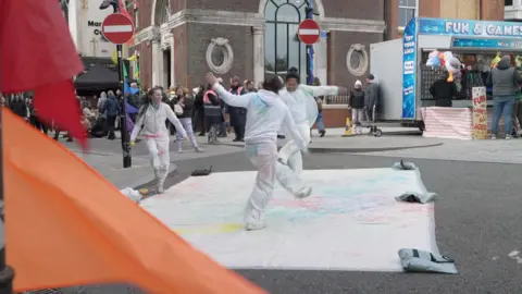 Three youths perform on a white mat that is weighted down in the middle of the road.  They wear white overalls with paint on them.
