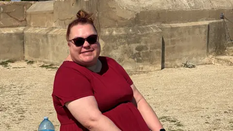 Michael Costello A woman in red t-shirt leans backwards on a beach wearing black sunglasses. 