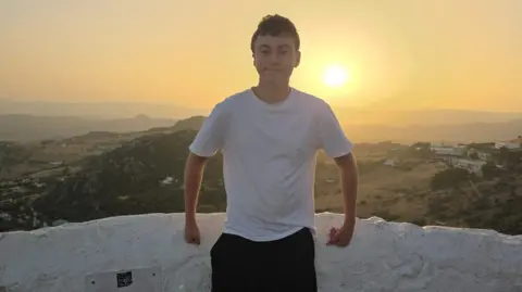 Will Will Payne smiles at the camera. He is stood in from of a sunset and hills. he is wearing 