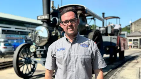 James Quirk is wearing a short sleeved grey shirt and glasses. He has short dark hair and is smiling standing in front of a large steam locomotive. It is a sunny day.