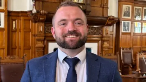 Emma Draper Devon smiles broadly for a photo inside a the council chamber. He has short brown hair and wears a navy suit with his arms behind his back.