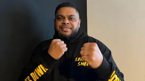 Shelim Ali An man with dark hair and a beard holds his fists up while smiling and wearing a black hoodie with gold-coloured lettering on it