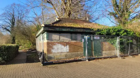 The fenced off toilet block pictured in March 2025. It is a one-storey brick house with green doors and matching window seals. It is surrounded by a metal fence. 