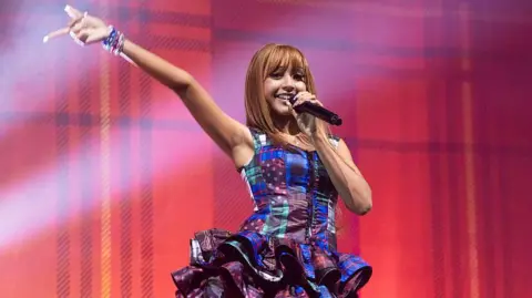 Getty Images PinkPantheress performs on stage wearing a ruffled blue dress with a green and black chequered design. She has long brown hair with a fringe and holds a black microphone 