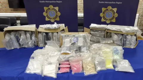 An Garda Síochána Bags of suspected drugs lined up on a table with posters of An Garda Síochána's logo hung up behind it