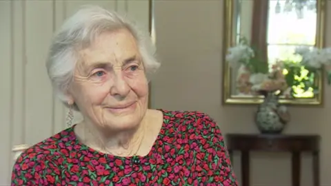 A 92-year-old woman with short grey hair and a red and pink flowery top smiles. She is sat in a well decorated room with a flower vase and mirror behind her.