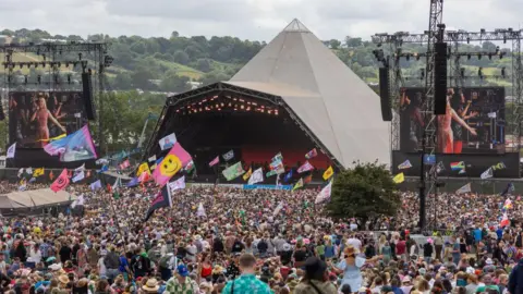 A huge crowed with lots of large flags assembled in front of the Glastonbury pyramid stage. 
