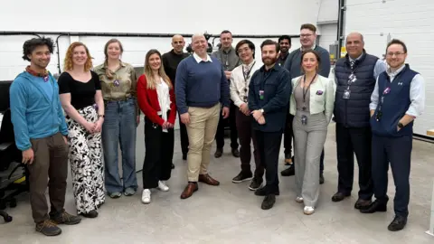 Fourteen people lining up for a group photo, smiling at the camera. They are the staff at Astral Systems, and are standing in what looks like a large warehouse building with the brick walls painted white. 