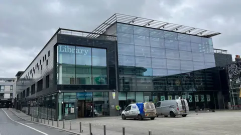 George Carden/BBC The Jubilee Library in Brighton which is a large 4 storeyed building with glass panels. It has a big library sign on the front and has a large pedestrian square at the front