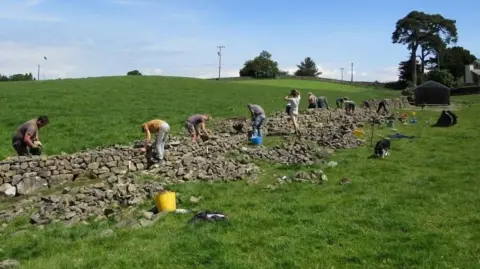 Mike Kitson Nine men and one women are working on building a drystone wall across two grassy fields. Some are lifting stones and others are placing stones on the wall. There are large piles of stones next to the wall.