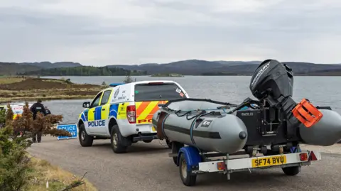Jasperimage Police pickup truck towing an inflatable boat on a trailer parked by a lakeside, with officers and another police vehicle in the background near a body of water and low hills under an overcast sky.