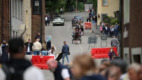 General image of horses pulling small two-wheel carriages down a hill in Appleby. A number of people watch on from the pavements.