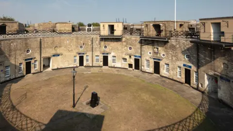 The fort, viewed from a first floor balcony. It is a semi-circle stone building with iron fencing around the top. There are doors and windows evenly spaced. The building is built around a circular lawn. There is a black lamppost in the middle of the lawn.