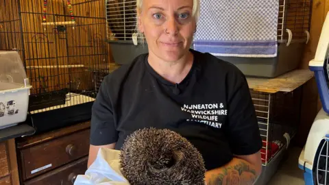 Emma Hudson of the Nuneaton and Warwickshire Wildlife Sanctuary holds a hedgehog in a blanket and looks towards the camera, with cages in the background