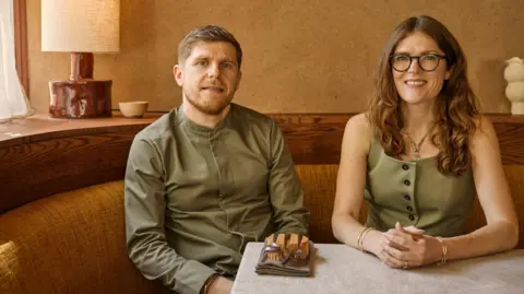 Safia Sarkarchi Kirk Haworth, a man with light hair and facial hair, wearing an olive green shirt, sitting alongside Keeley Haworth, a woman with long wavy ginger hair, glasses, wearing an olive green dress.