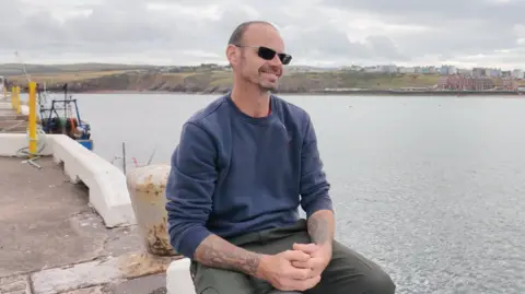 Nicky Lewis: a smiling man with very short, dark hair and a short beard, seated on the harbour front. He is wearing dark glasses, a dark blue sweatshirt and grey trousers. He has tattoes visible on both arms.