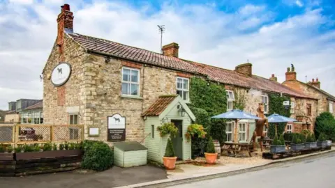 Planning Documents A traditional stone pub called the Black Horse Inn, with a red tiled roof, ivy on the walls, blue umbrella outdoor seating and a large horse statue beside the tables.