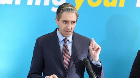 PA Media Simon Harris, with greying dark hair, wearing in a dark suit, with his hand raised to emphasise a point while speaking at a podium